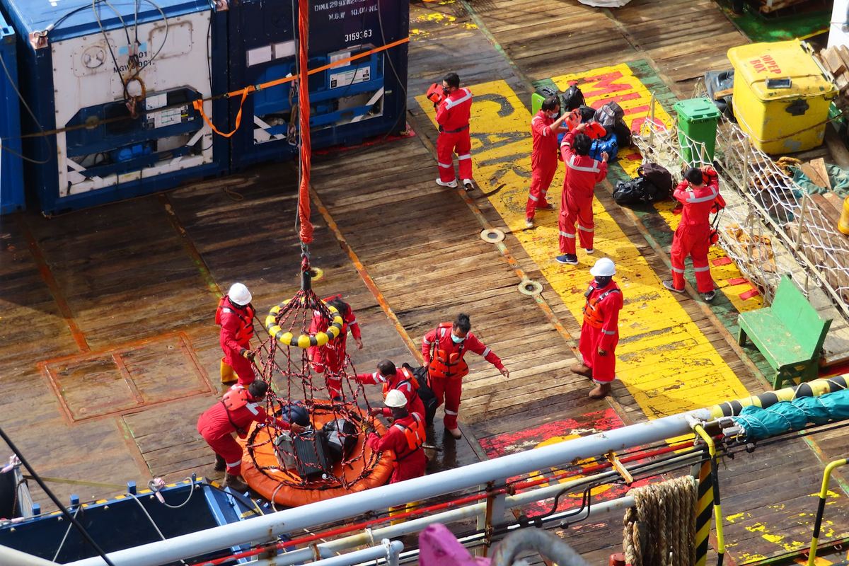 Offshore workers in red overalls on a platform deck as a crane lifts a crew basket